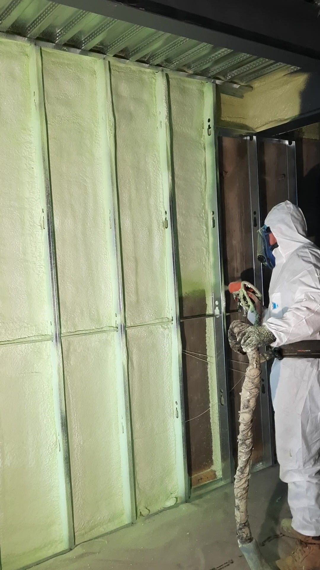 Une personne en combinaison de protection pulvérise de la mousse isolante sur un mur d'un bâtiment.