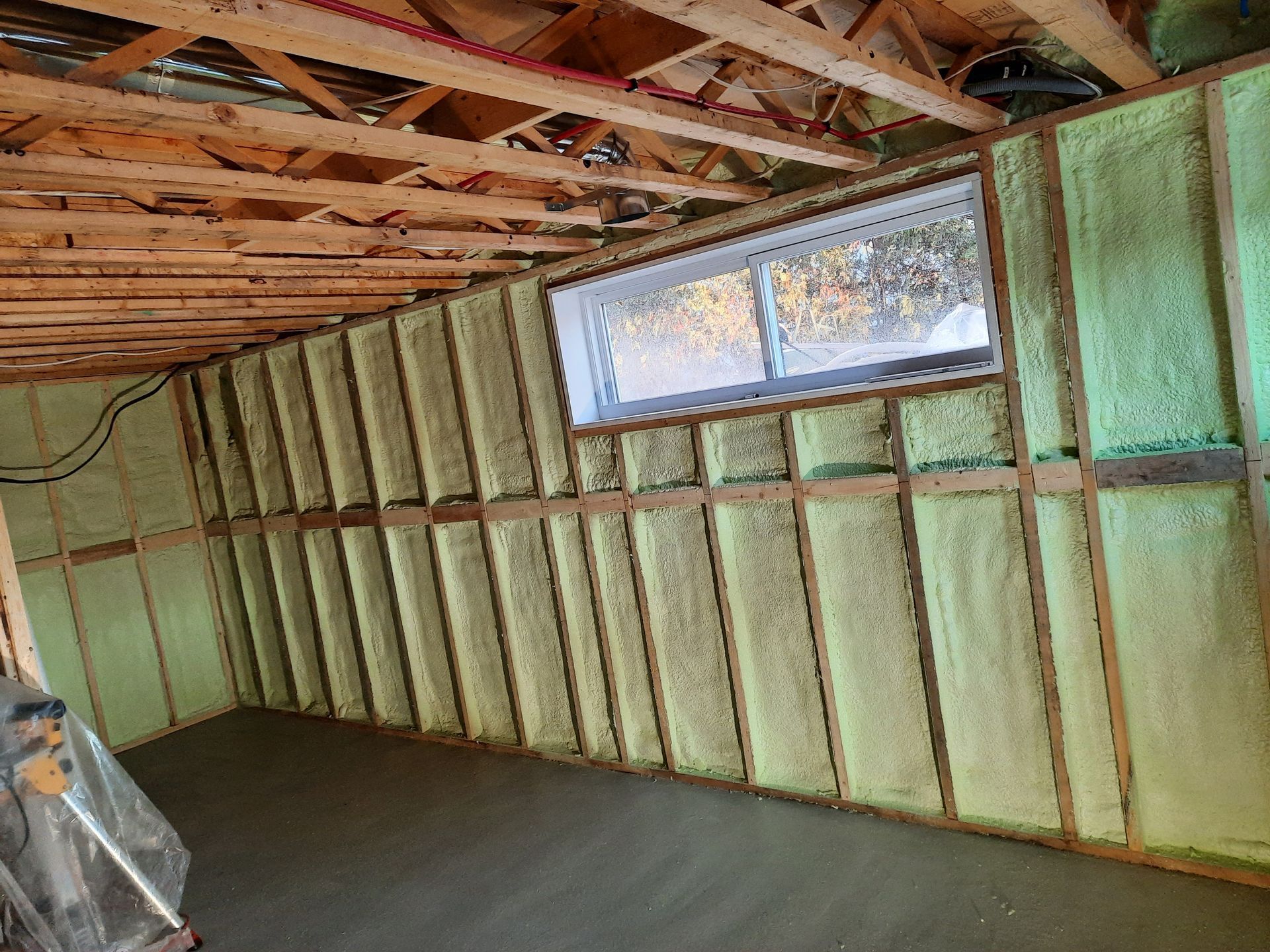 Mur de sous-sol avec isolation en mousse pulvérisée, fenêtre, charpente en bois apparente et plancher en béton.