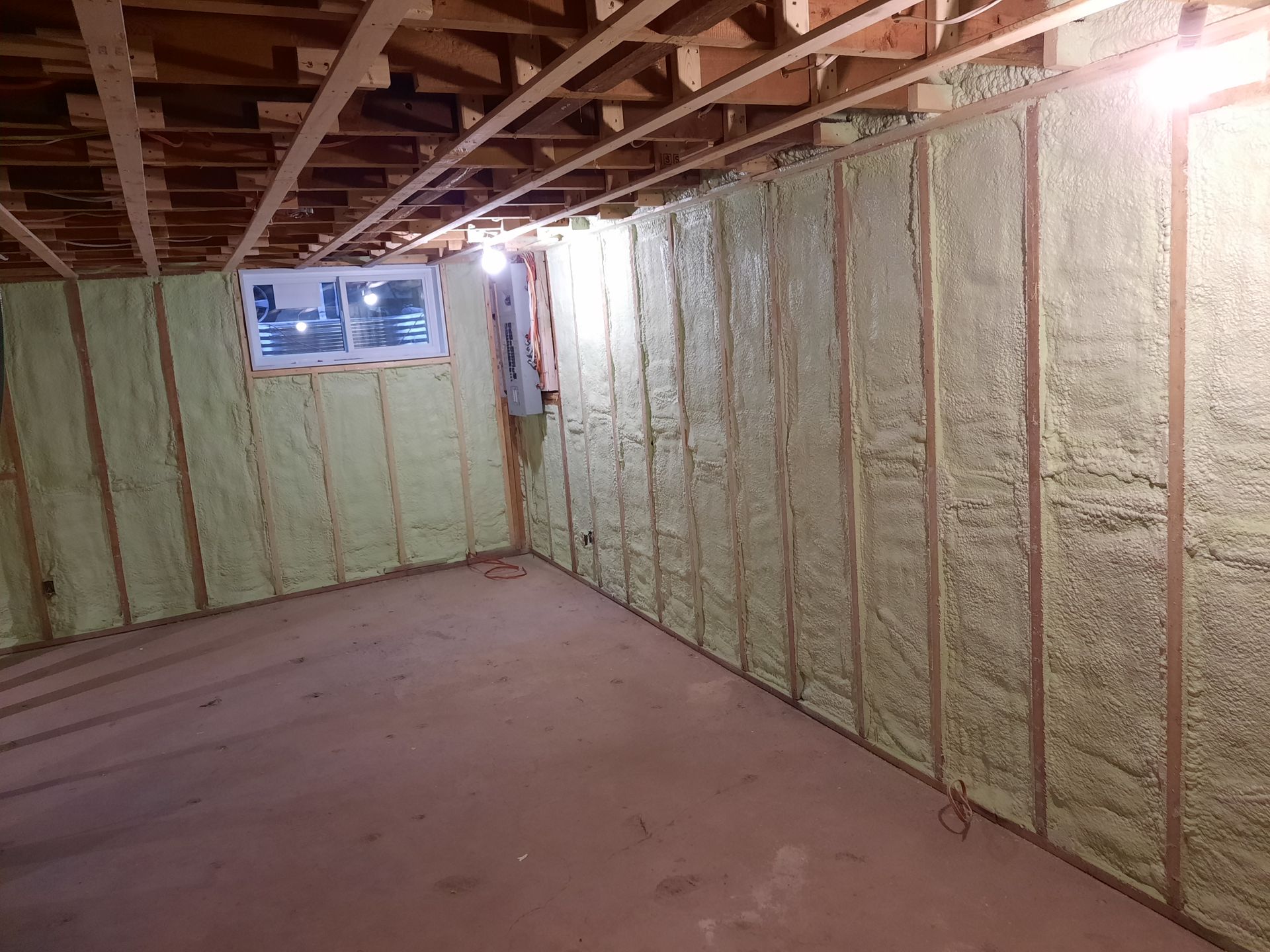 Sous-sol avec poutres apparentes en bois, murs recouverts d'isolant en mousse pulvérisée jaune et une petite fenêtre.