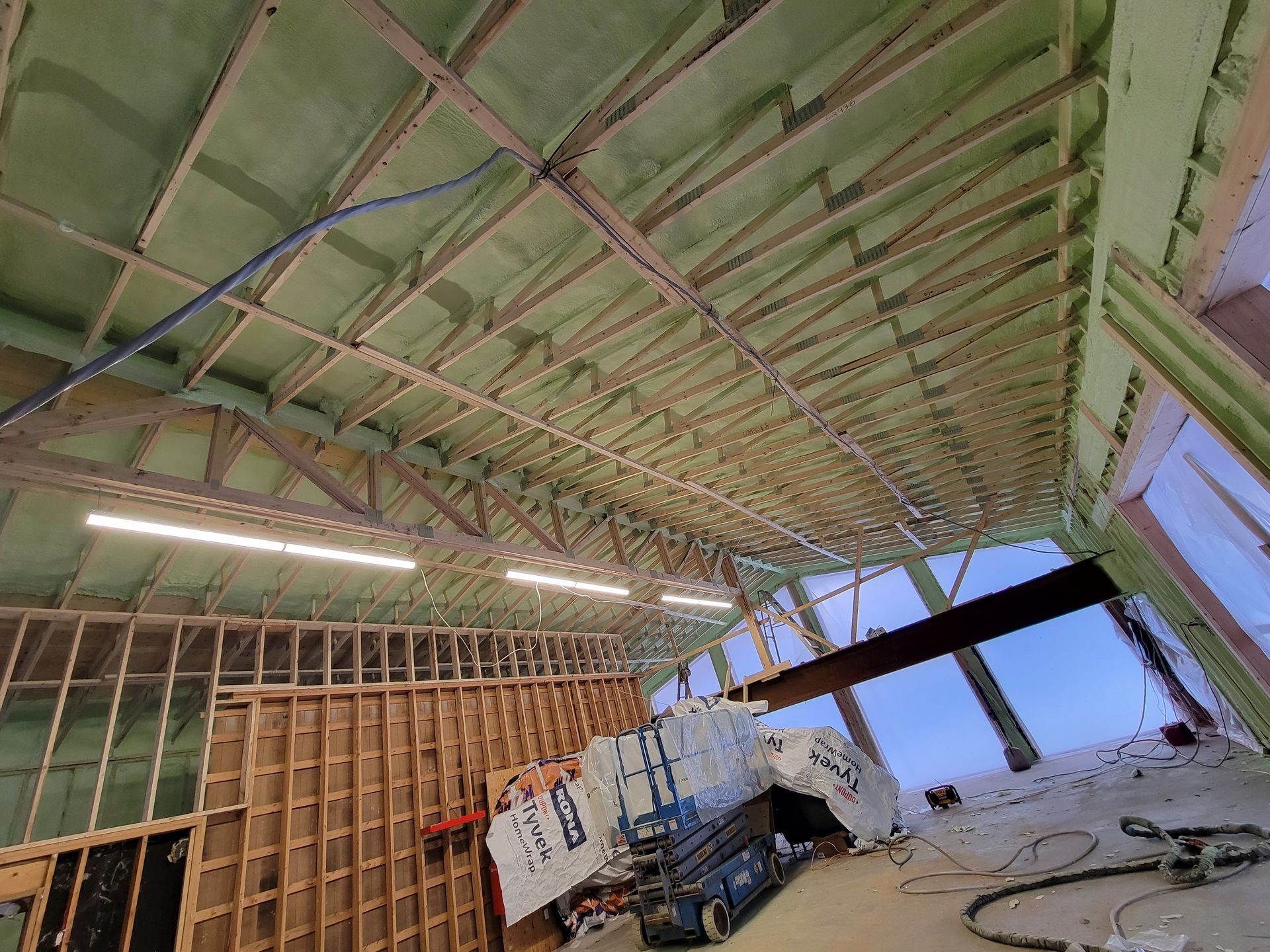 Intérieur d'un bâtiment en construction, avec isolation en mousse pulvérisée au plafond et charpente en bois visible.