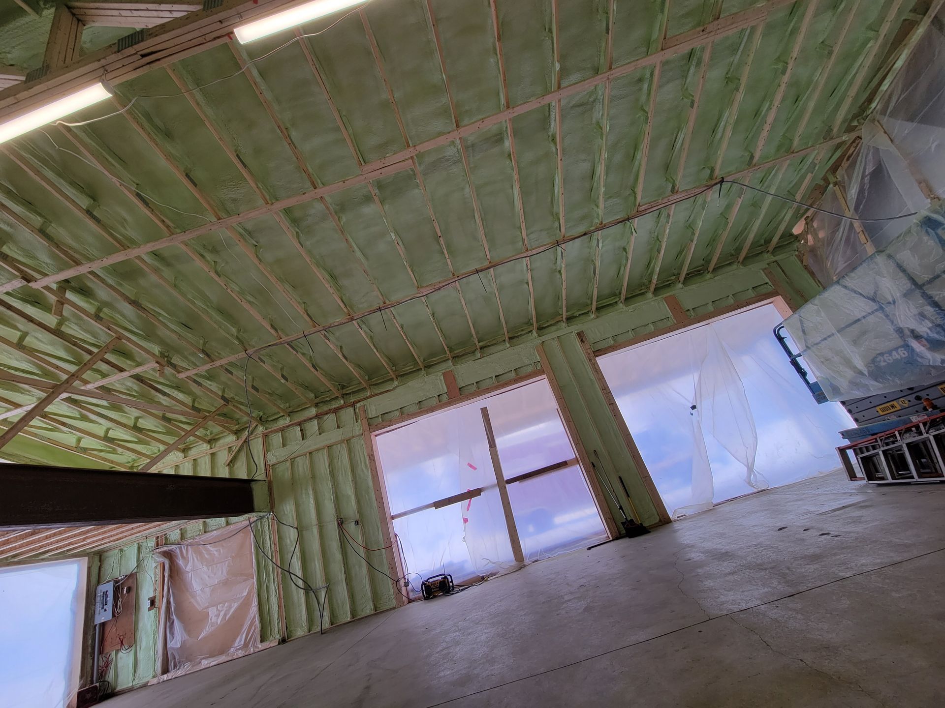 Vue intérieure d'un bâtiment en construction, montrant une isolation verte sur les murs et le plafond, les fenêtres et un sol en béton.