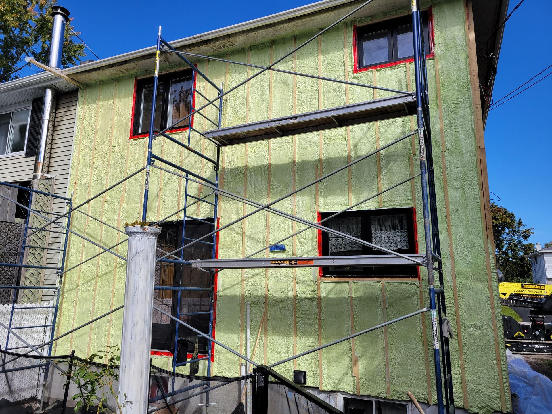 Extérieur de la maison avec isolation installée, échafaudage, fenêtres et ciel bleu.