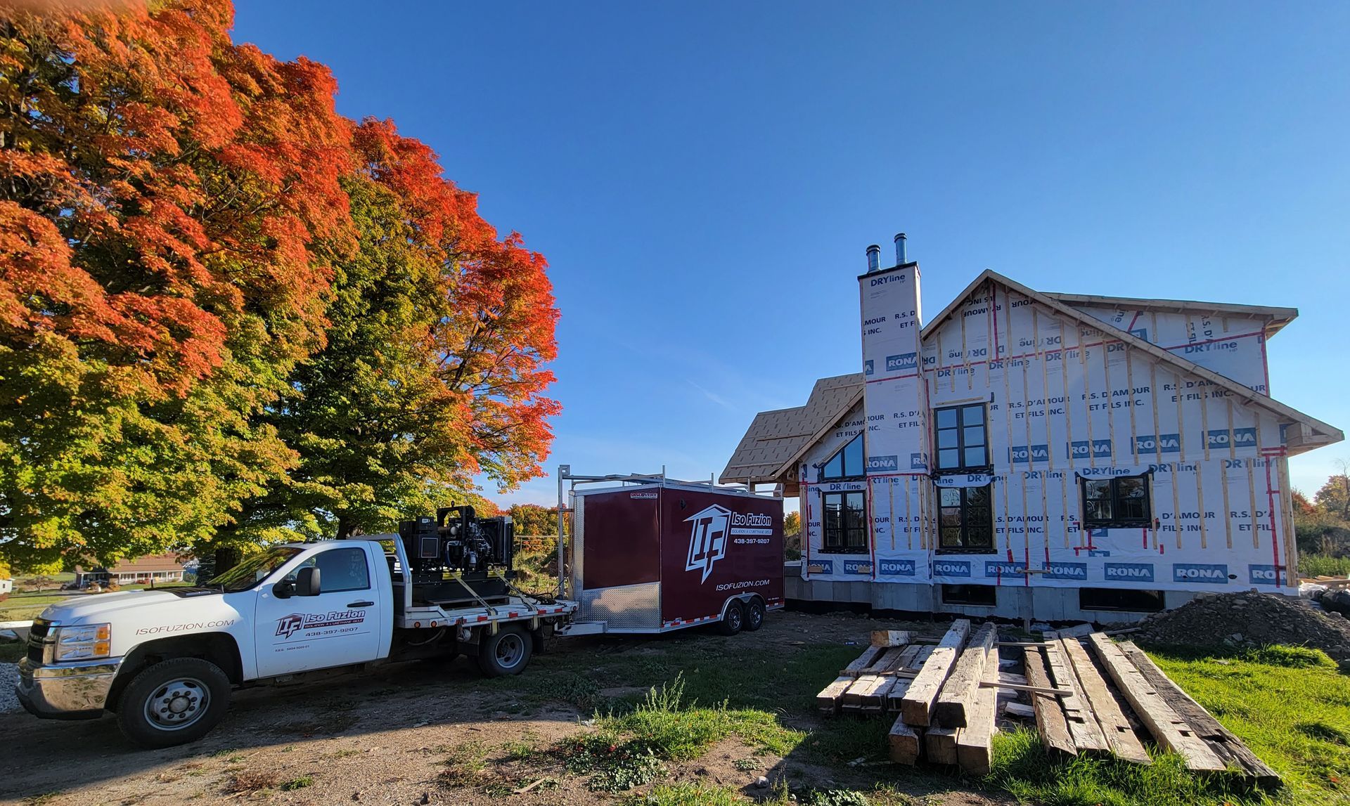 Chantier de construction : camion et remorque près d'une maison en construction, avec un feuillage d'automne coloré et un ciel bleu clair.