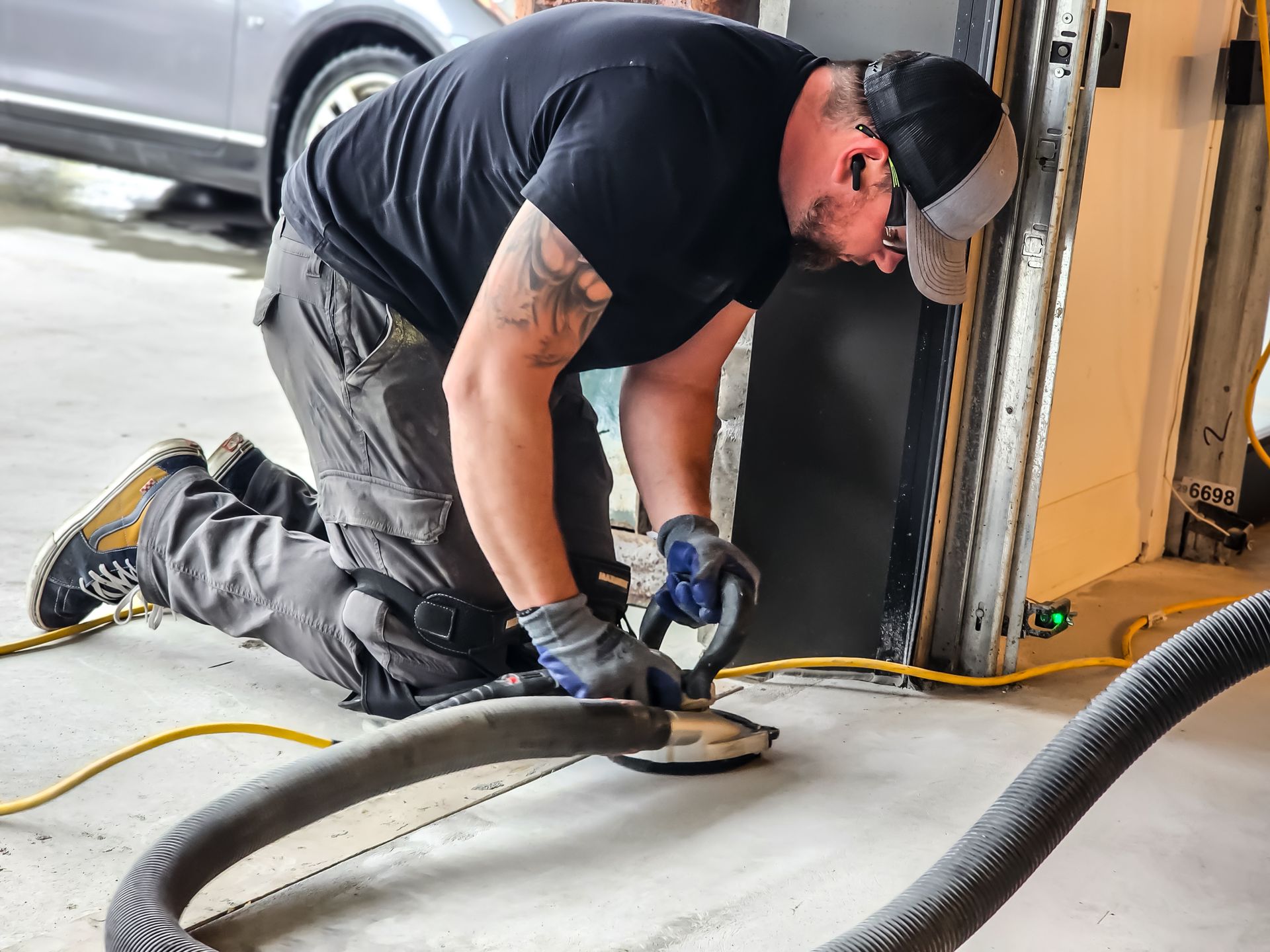Man kneeling, working with a hose and tool near a garage door. He wears gloves, a hat, and work clothes.