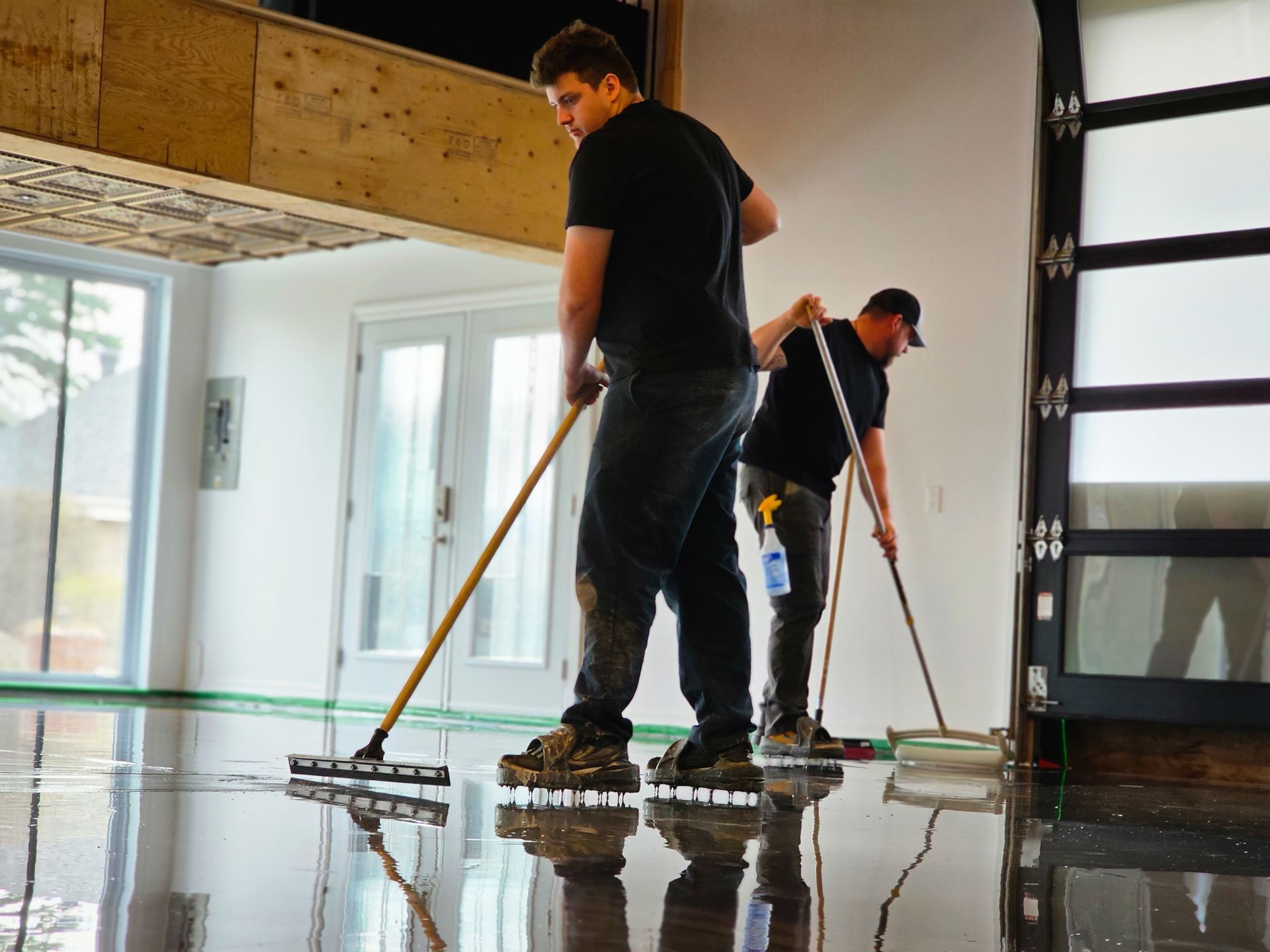 Two men applying flooring epoxy in a light-filled room with glass doors and windows.