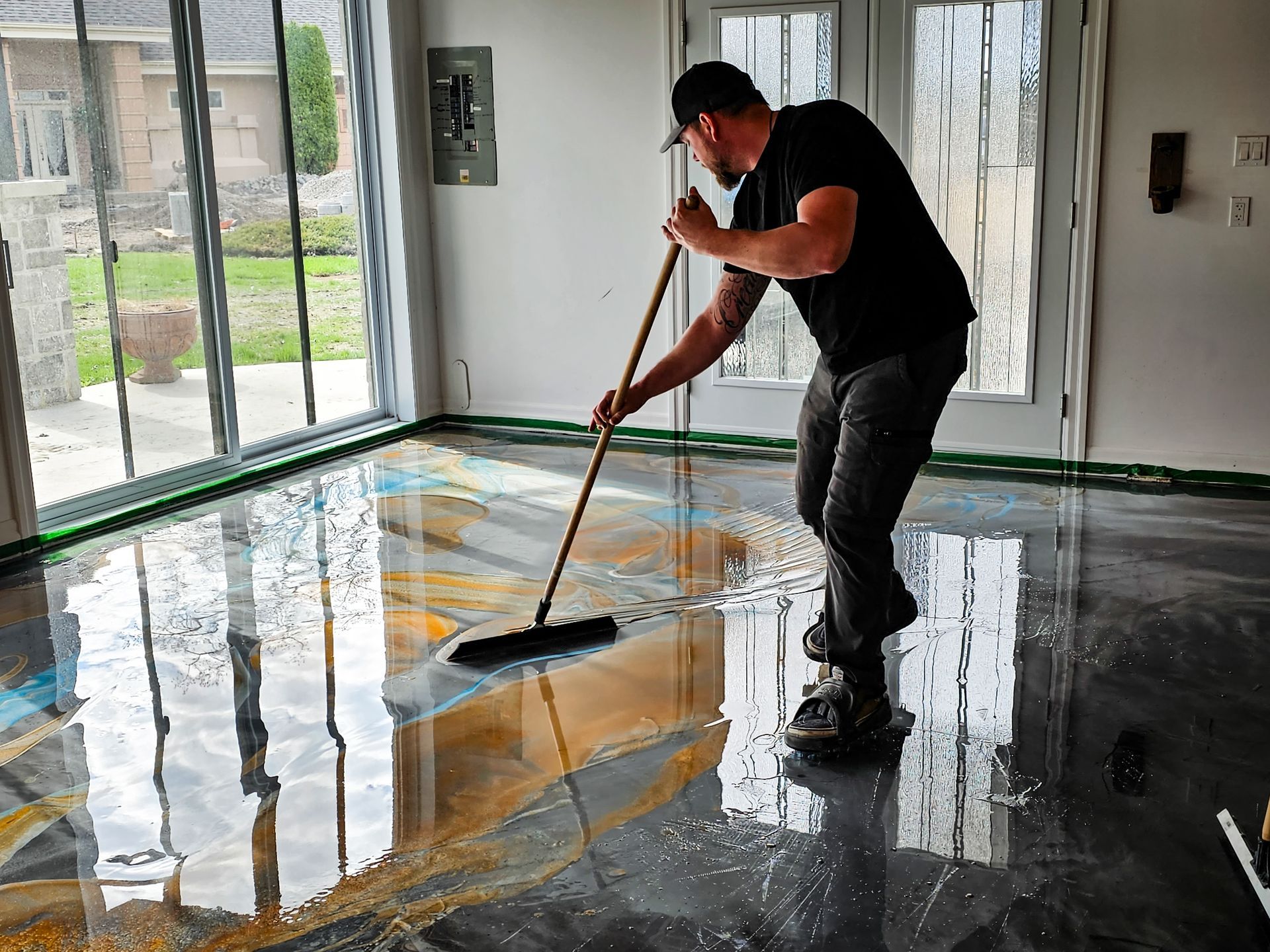 Man applying epoxy floor coating with a squeegee in a garage, mixing gold, blue, and gray colors.