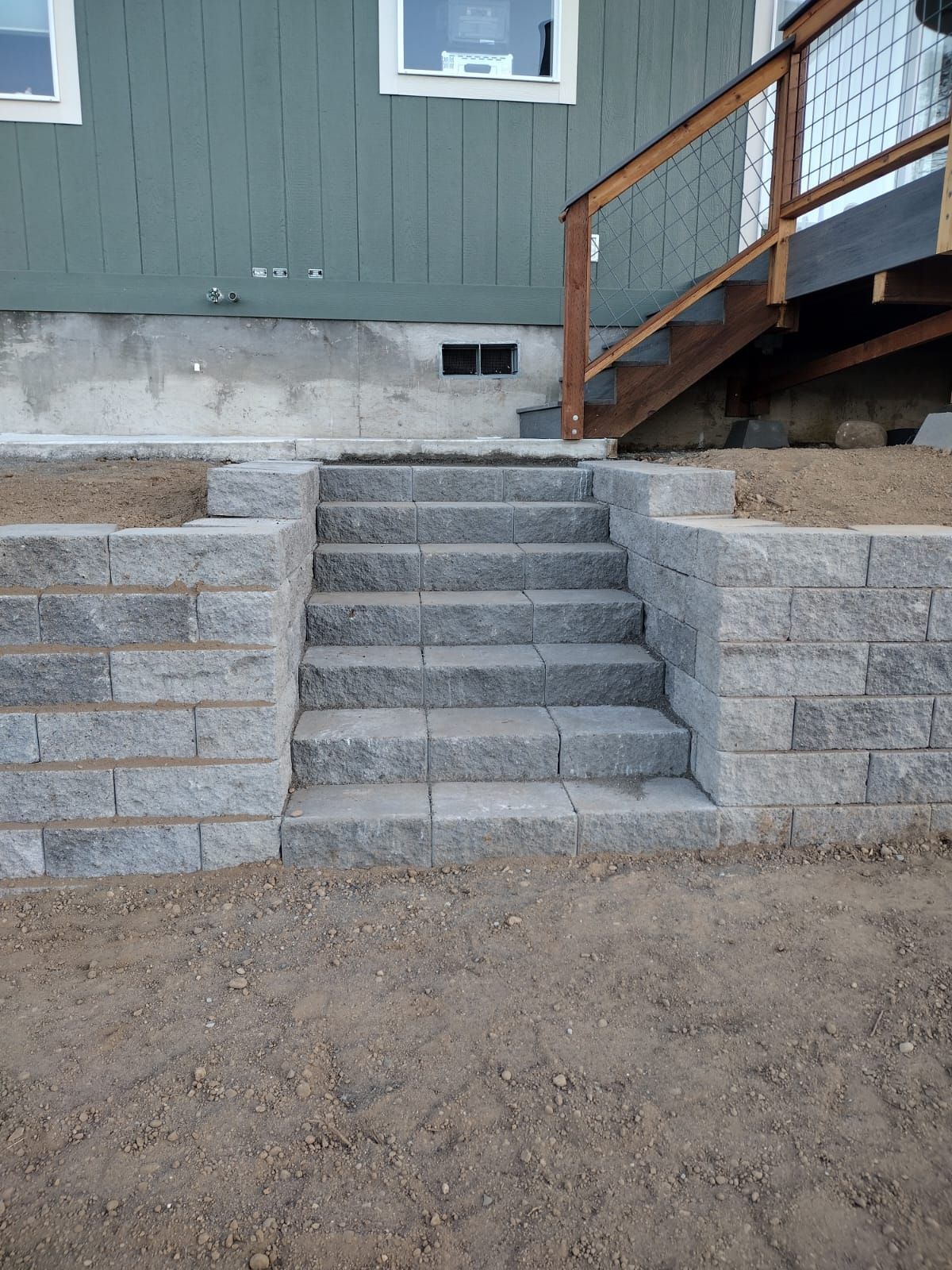 Stone steps leading up to a wooden deck, built into a retaining wall, with a green house exterior in the background.