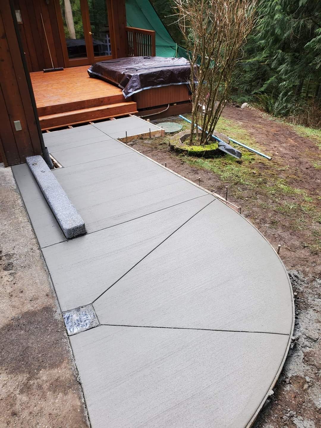 Newly poured concrete walkway with wooden deck in the background, set in a yard.