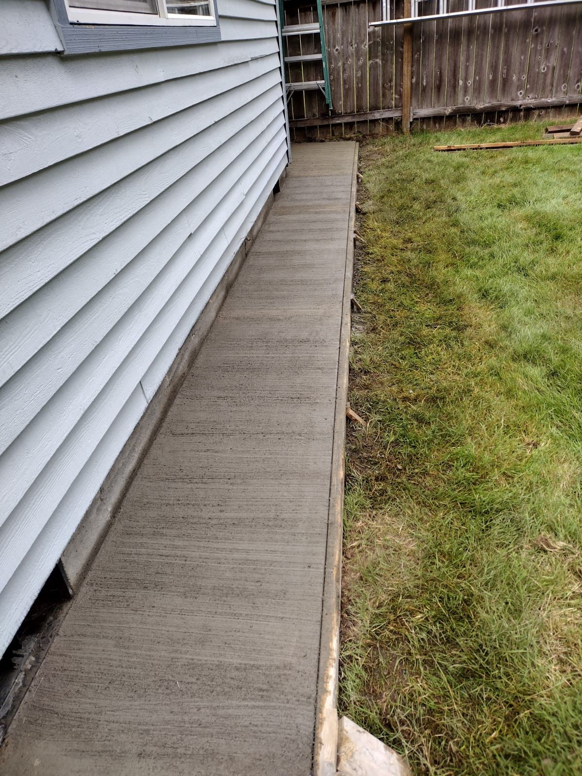 Newly poured concrete walkway along a light blue-sided house, next to a grassy lawn.