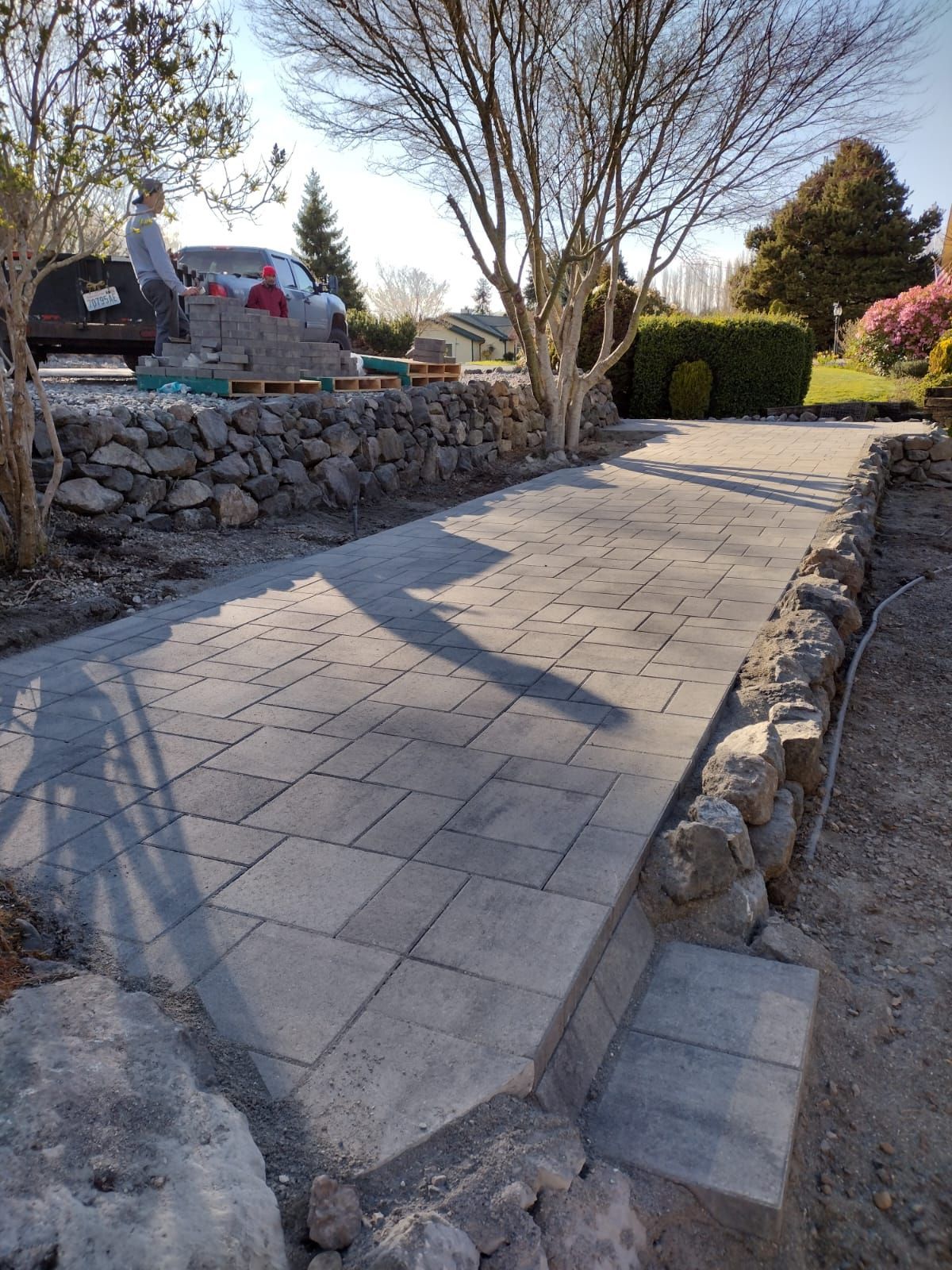 Stone paver pathway with rock retaining walls, leading to a yard with a person and construction materials.
