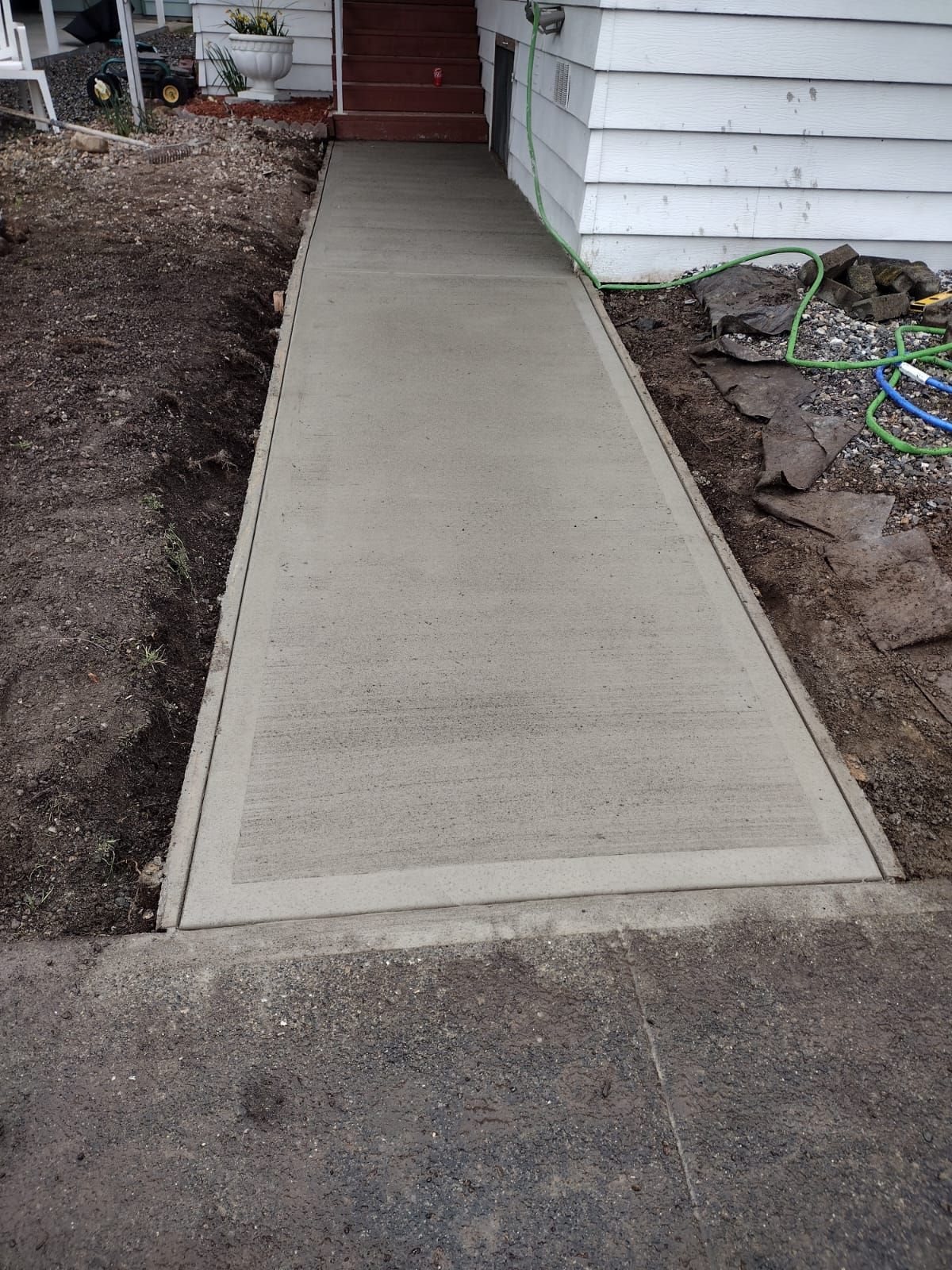 Concrete walkway leading to a house entrance. The path is grey and lined with soil.