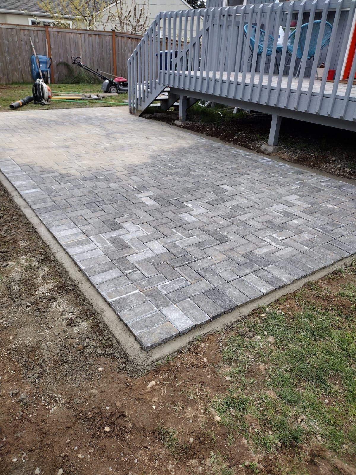 Brick patio in backyard, bordered by concrete, near a wooden deck.
