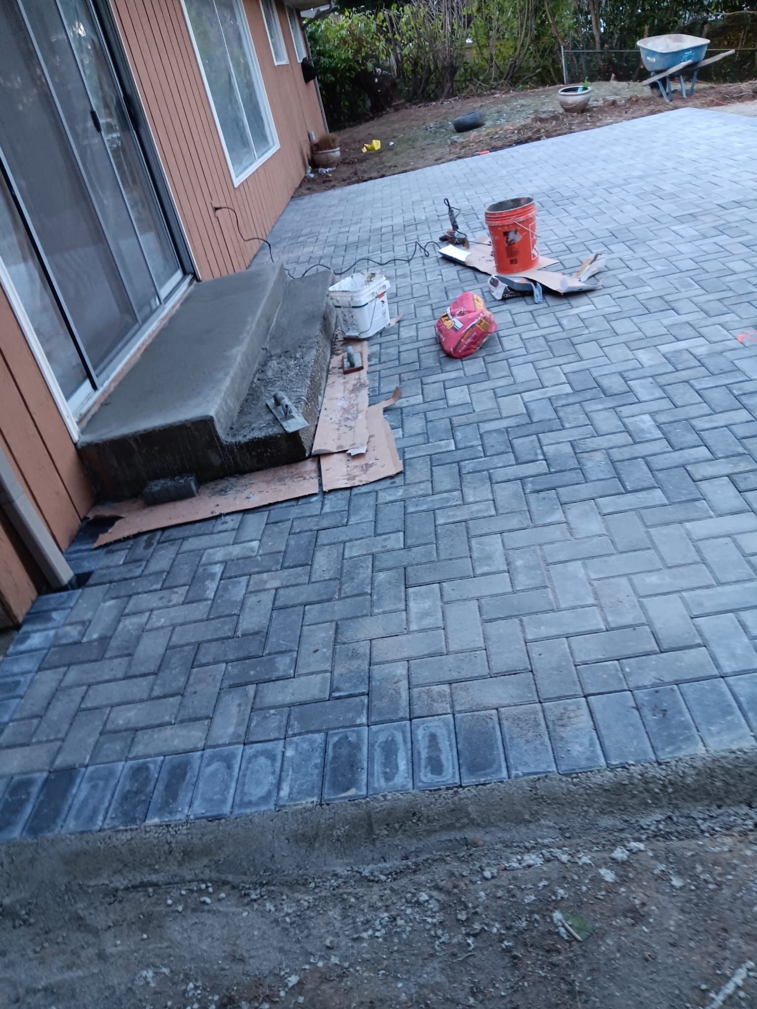 Brick patio construction outside a building with a concrete step, tools, and a red bucket visible.