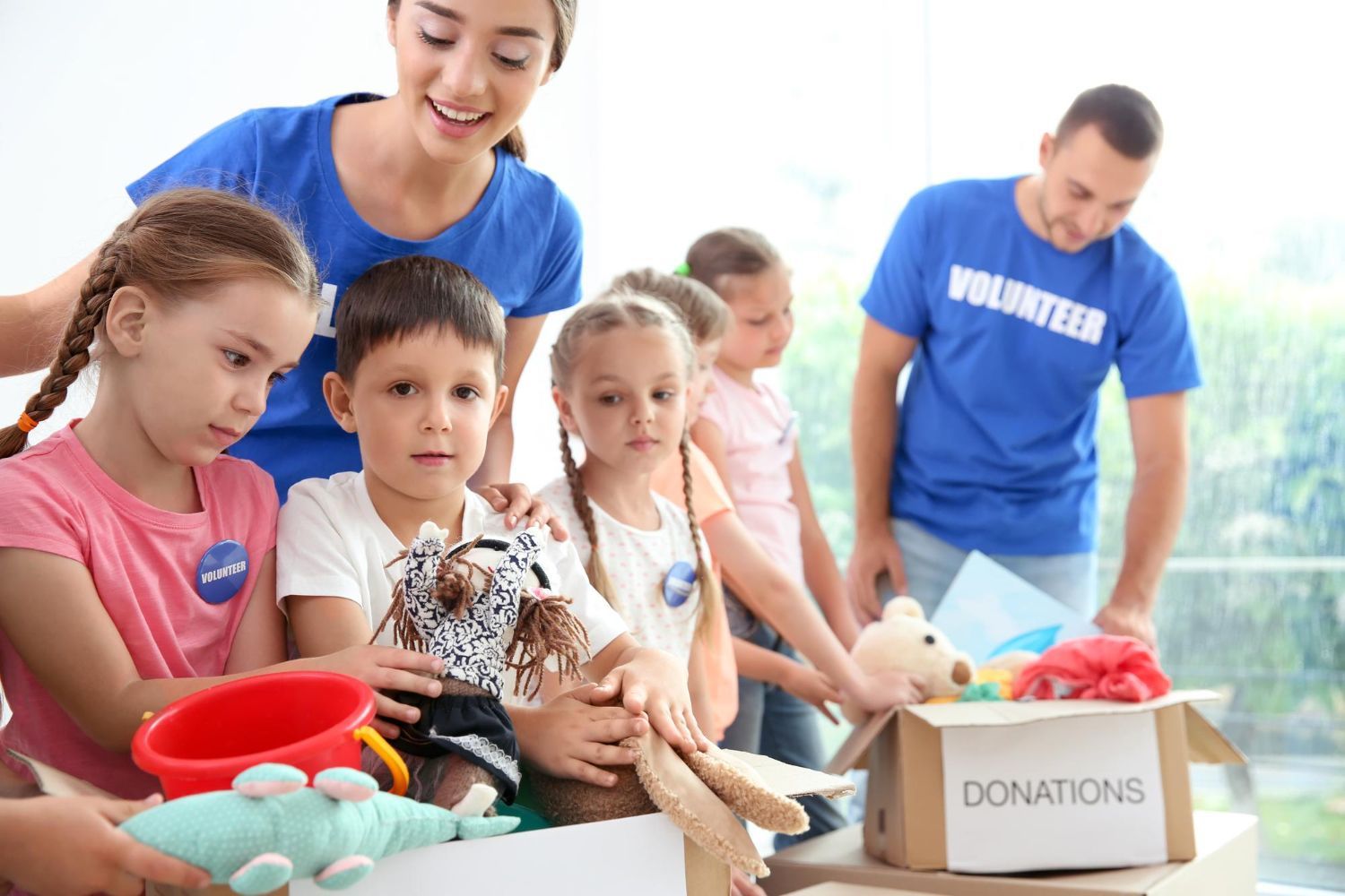 Volunteers and children sorting donations from a box. Children smile as they examine items; adults in blue shirts.