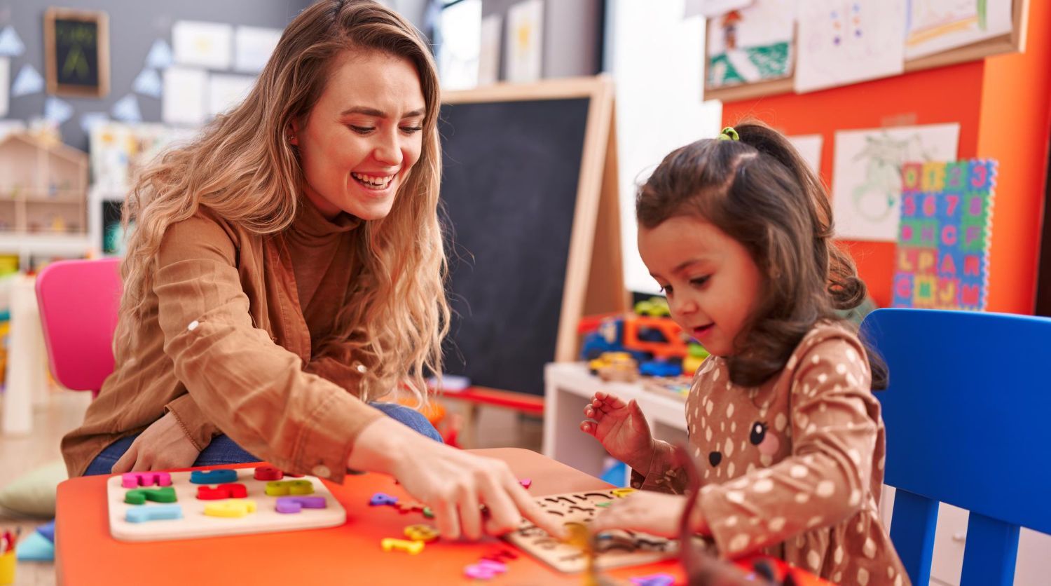 Teacher and young girl playing with colorful shapes at a table in a classroom.