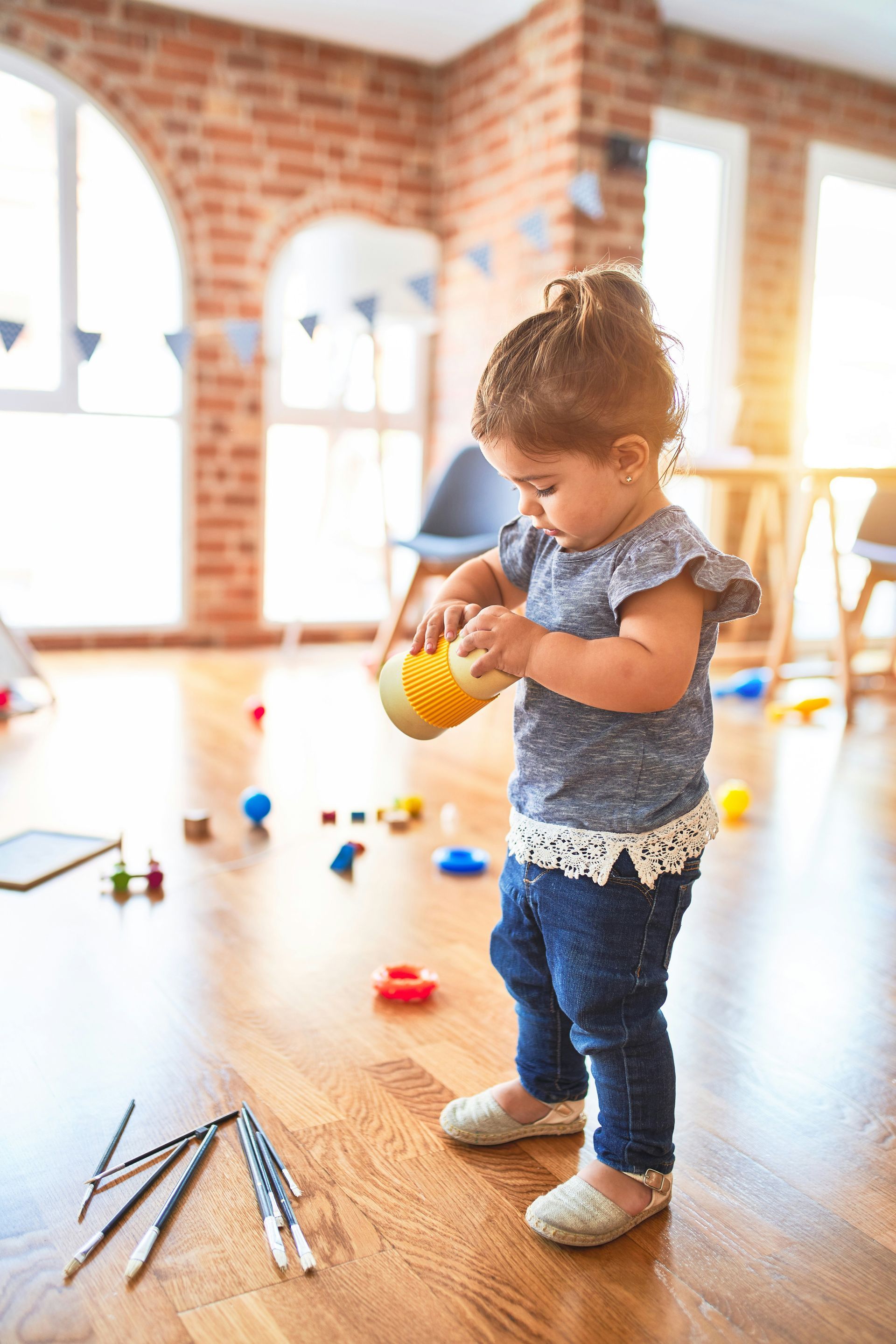 Young child playing with a toy in a brightly lit room with wooden floors and toys scattered.