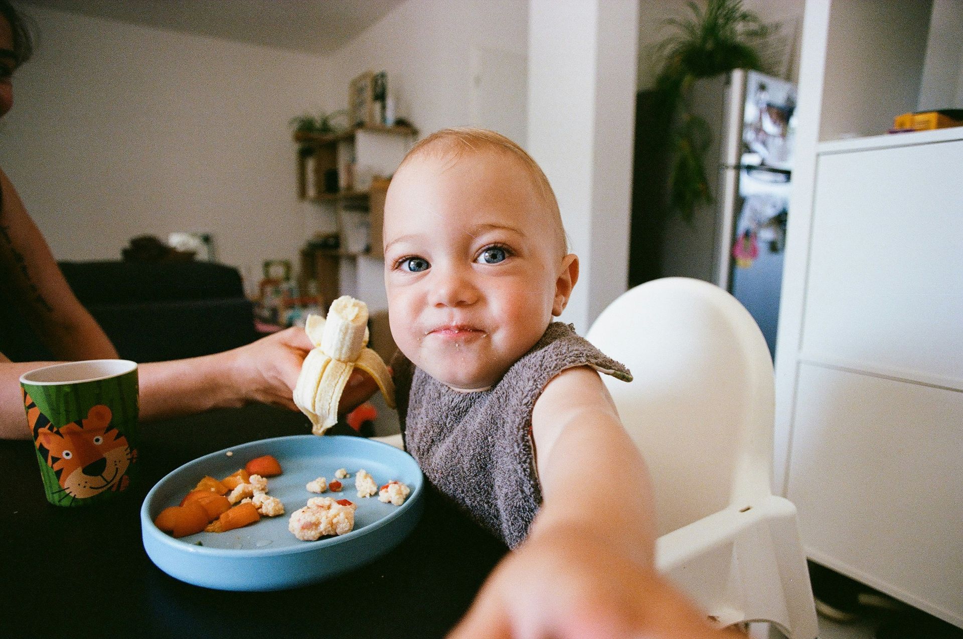 Smiling baby in high chair eating banana, carrots, and other food at a table.