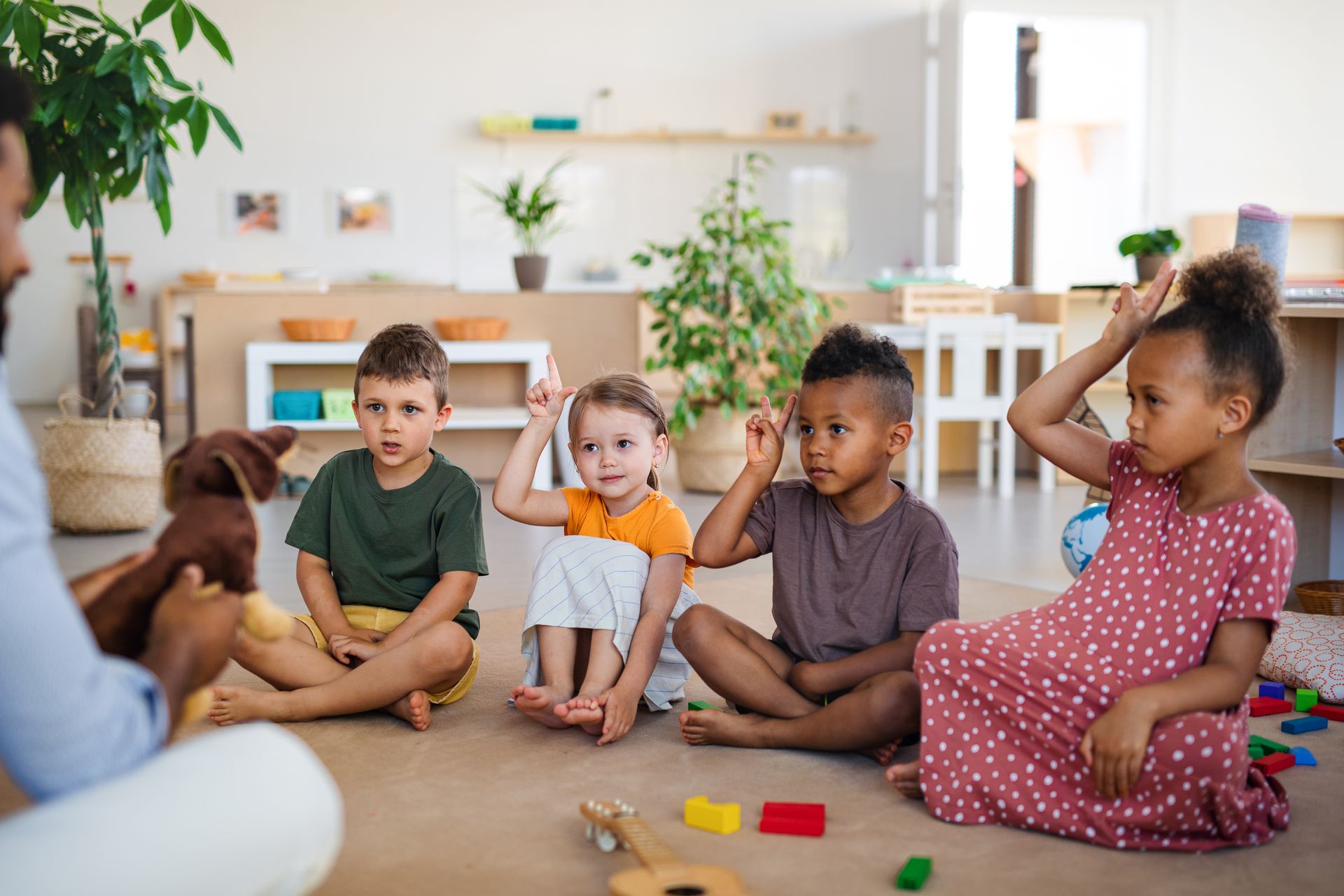 A teacher with a puppet interacts with four children seated on the floor, raising their hands.