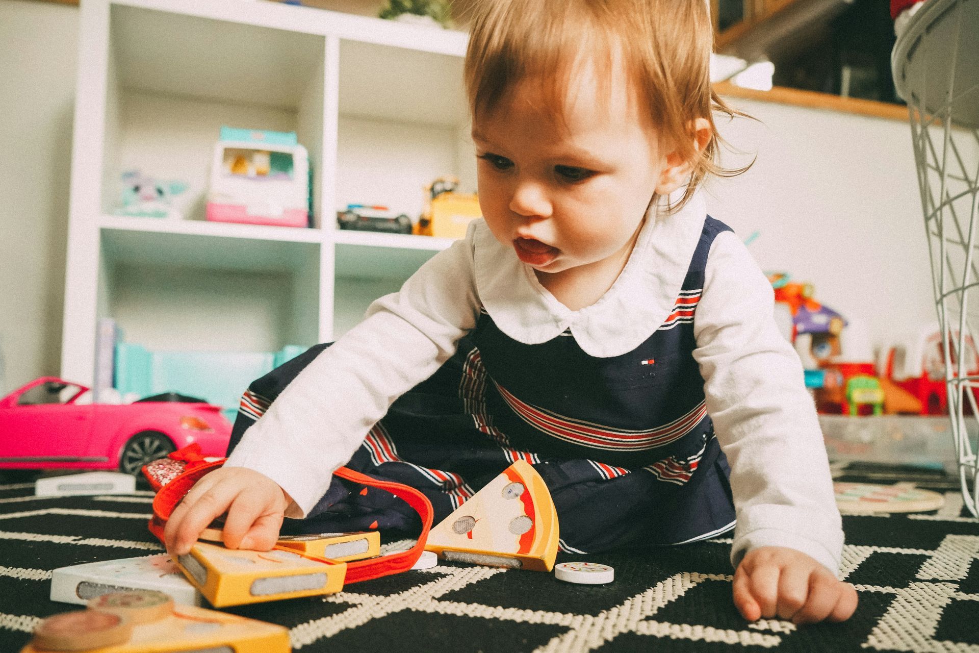Young child plays with toy food on a patterned rug, near a shelf filled with toys.