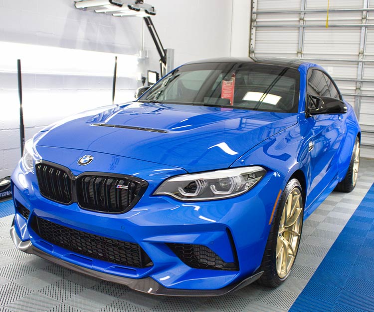 Blue BMW M2 sports car with gold rims, parked indoors with detailing lights.