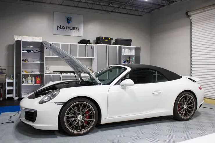 A white porsche 911 cabriolet is parked in a garage with its hood up.