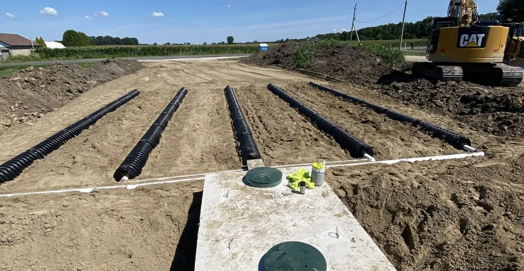 A cat excavator is driving down a dirt road next to a septic tank.