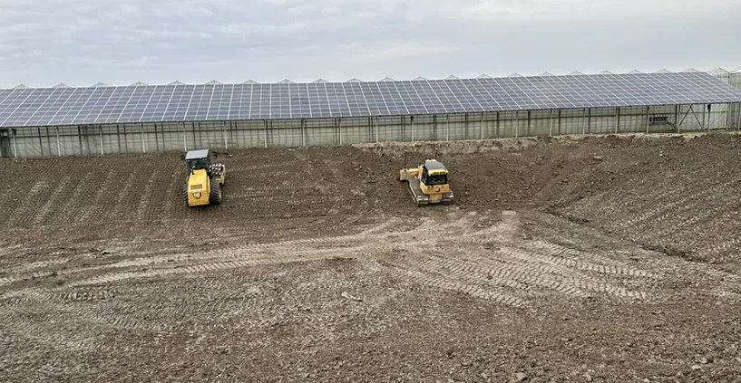 Two tractors are working in a field with solar panels in the background.