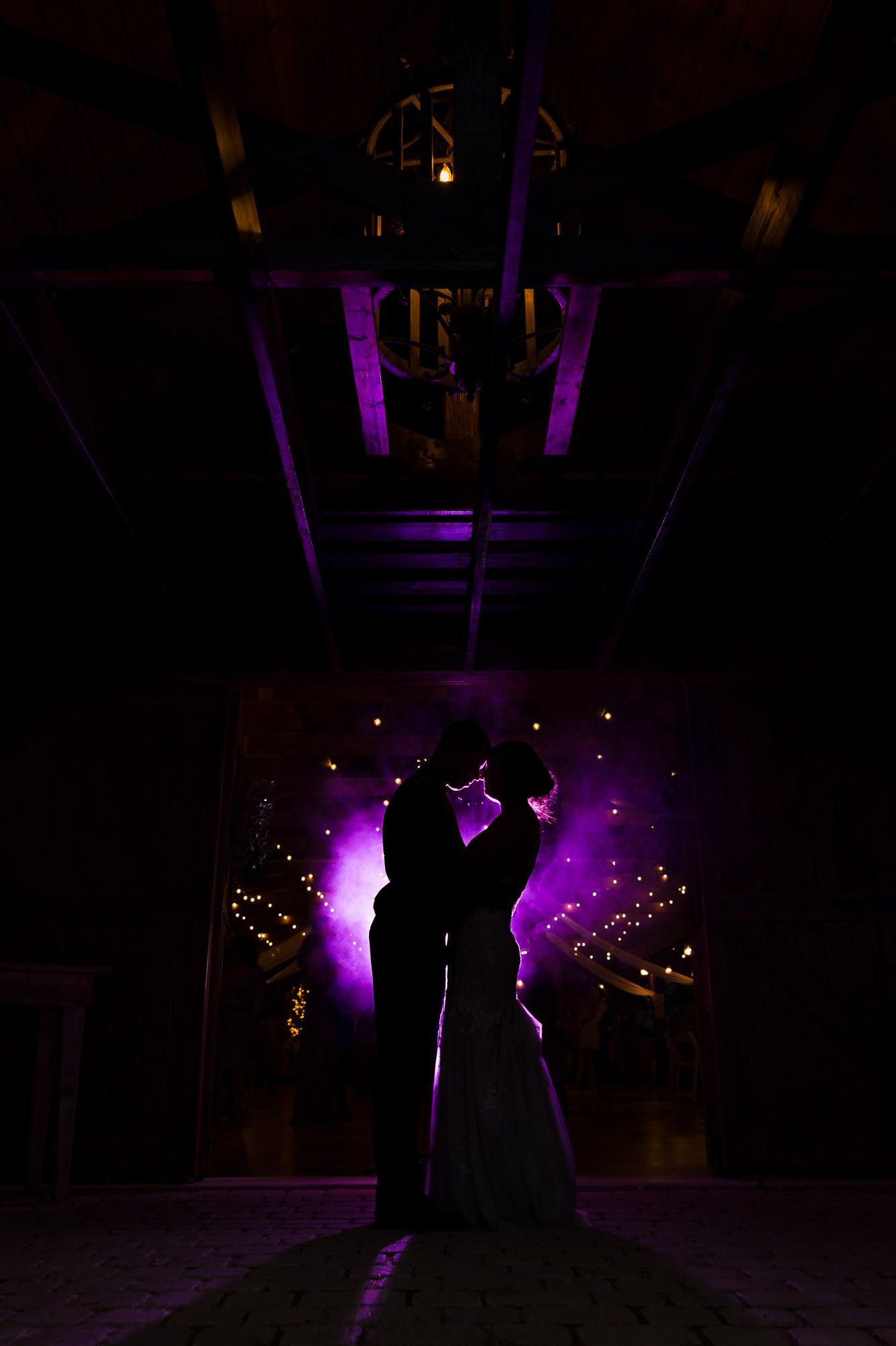A bride and groom are dancing in a dark room with purple lights behind them.