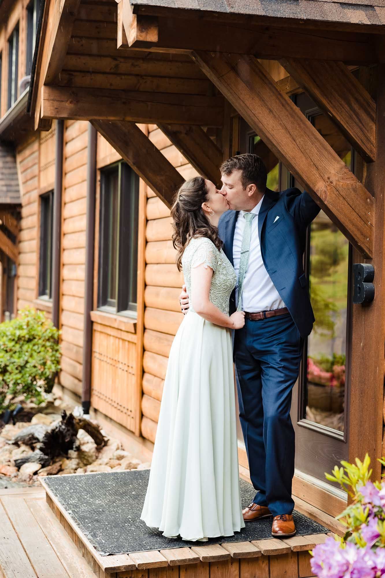 A bride and groom are kissing in front of a log cabin.