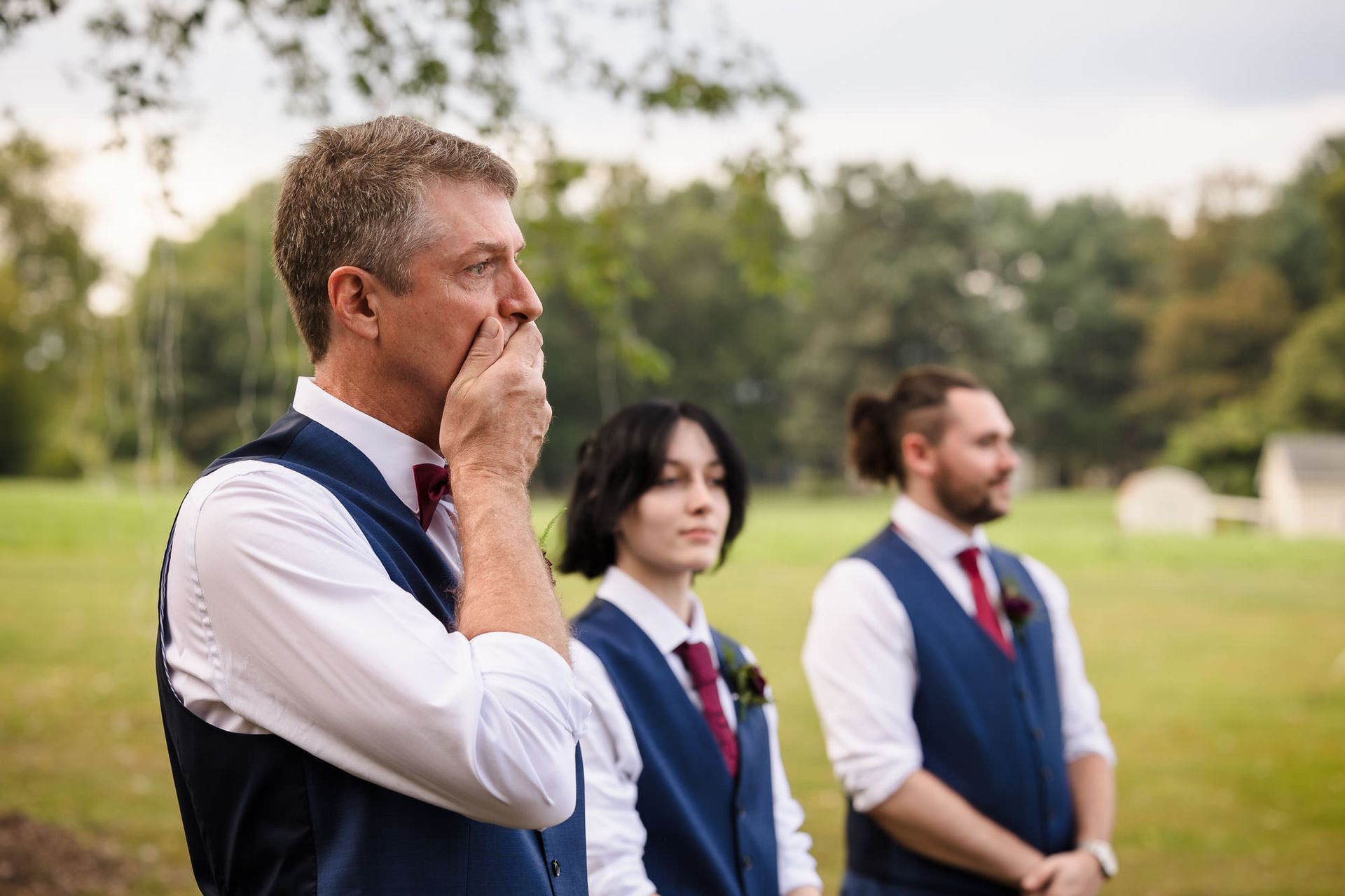 A man is covering his mouth while watching a bride and groom walk down the aisle at a wedding.