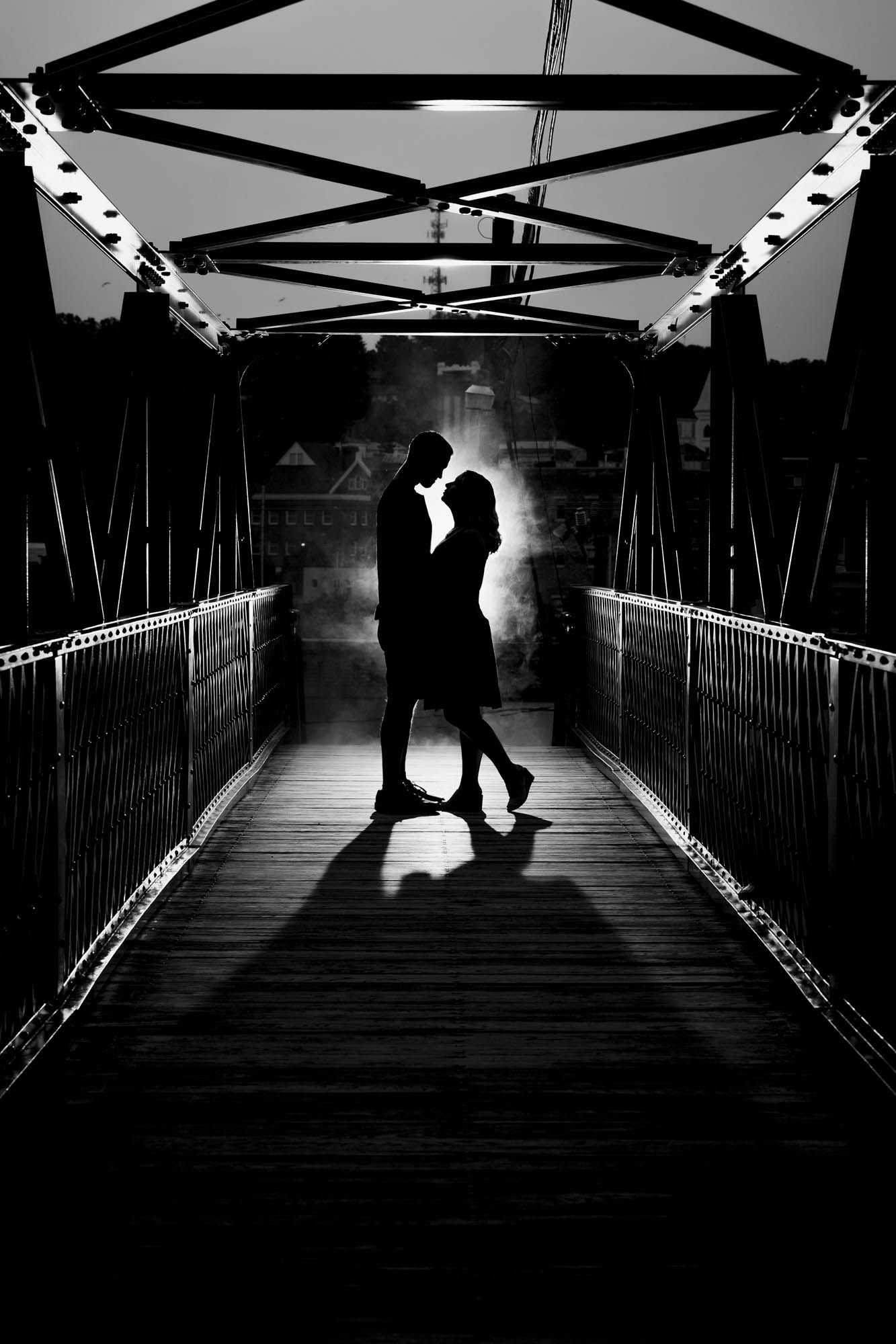 A black and white photo of a man and woman kissing on a bridge.