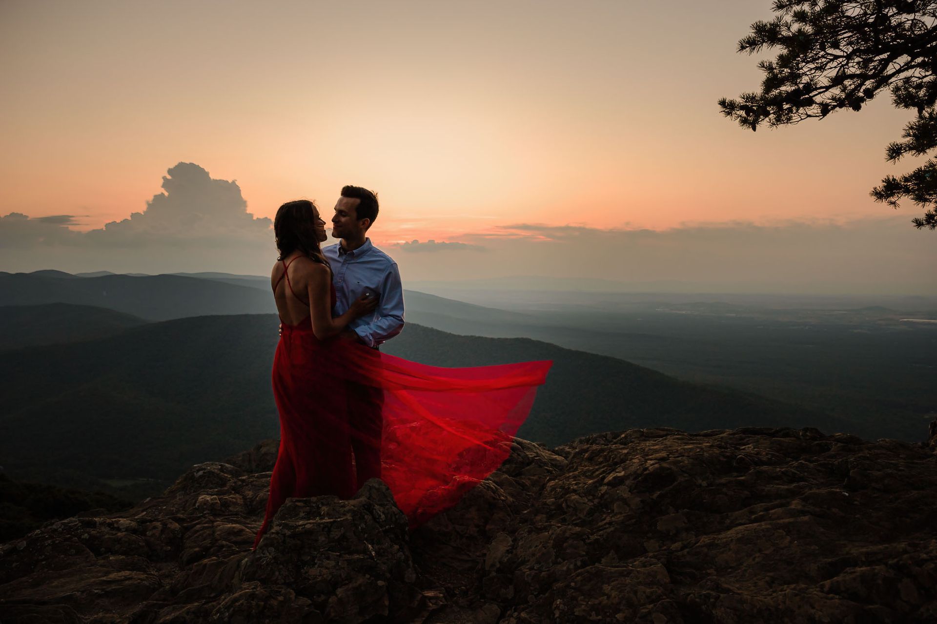 A man and a woman are standing on top of a mountain at sunset.