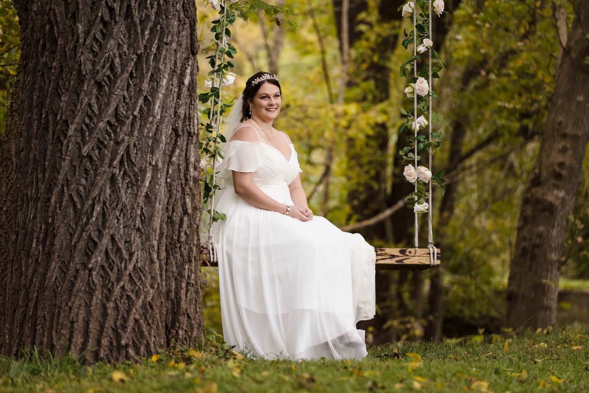 A bride in a wedding dress is sitting on a swing under a tree.