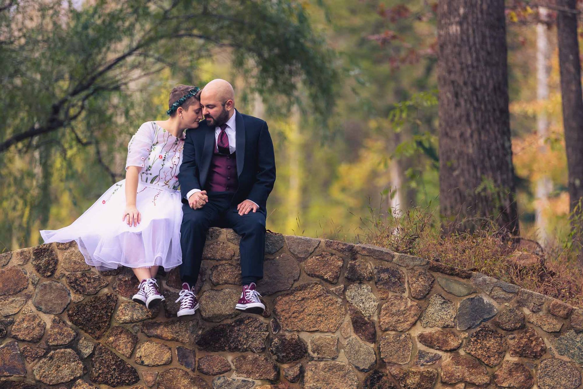 A bride and groom are sitting on a stone wall in the woods.