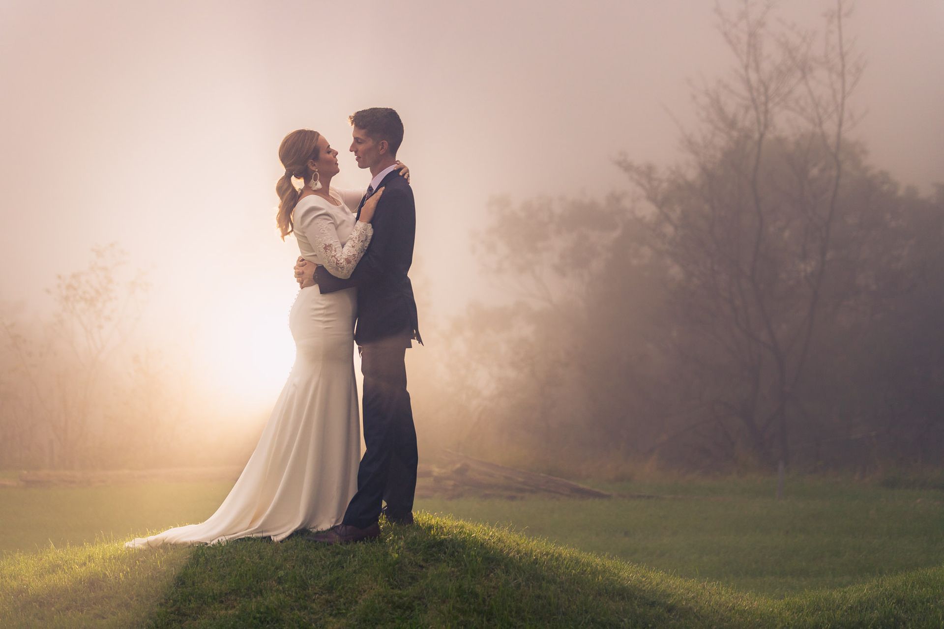 A bride and groom are standing on top of a grassy hill in the fog.