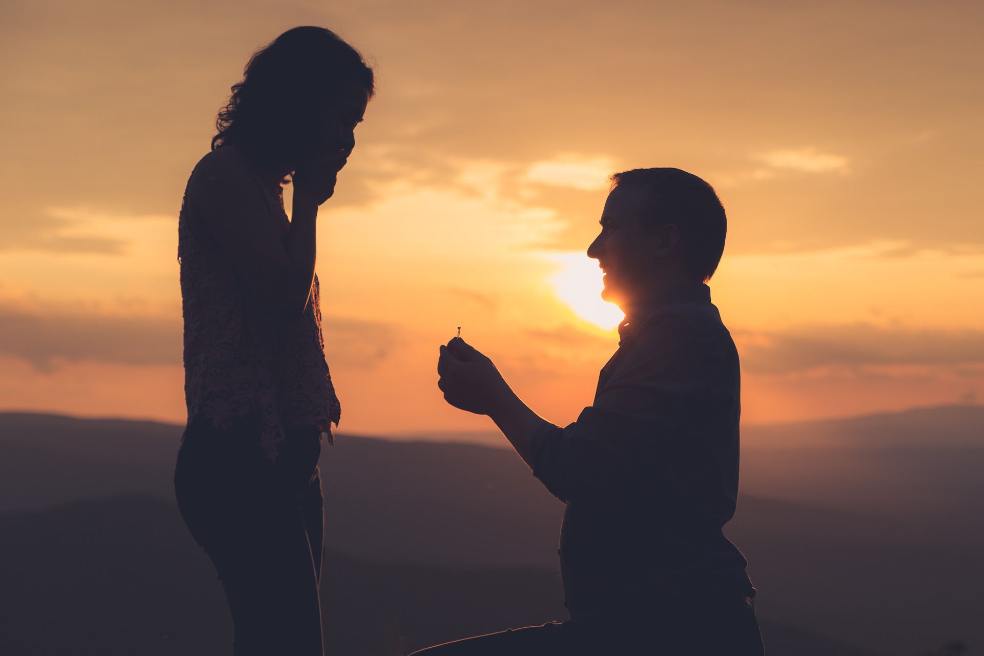 A man is kneeling down to propose to a woman at sunset.