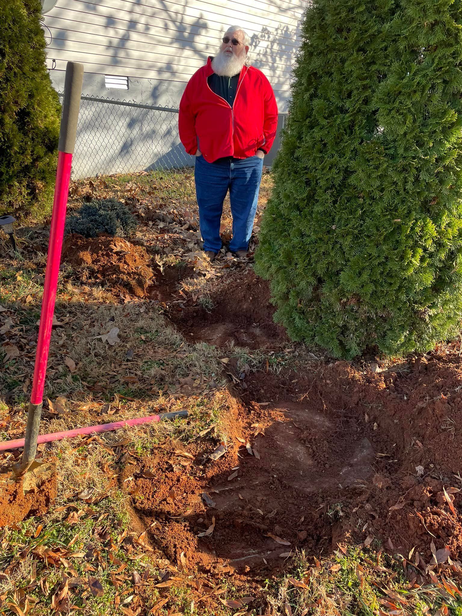 One of the client in a red jacket is standing in the hole ready for his new septic systm next to a tree.