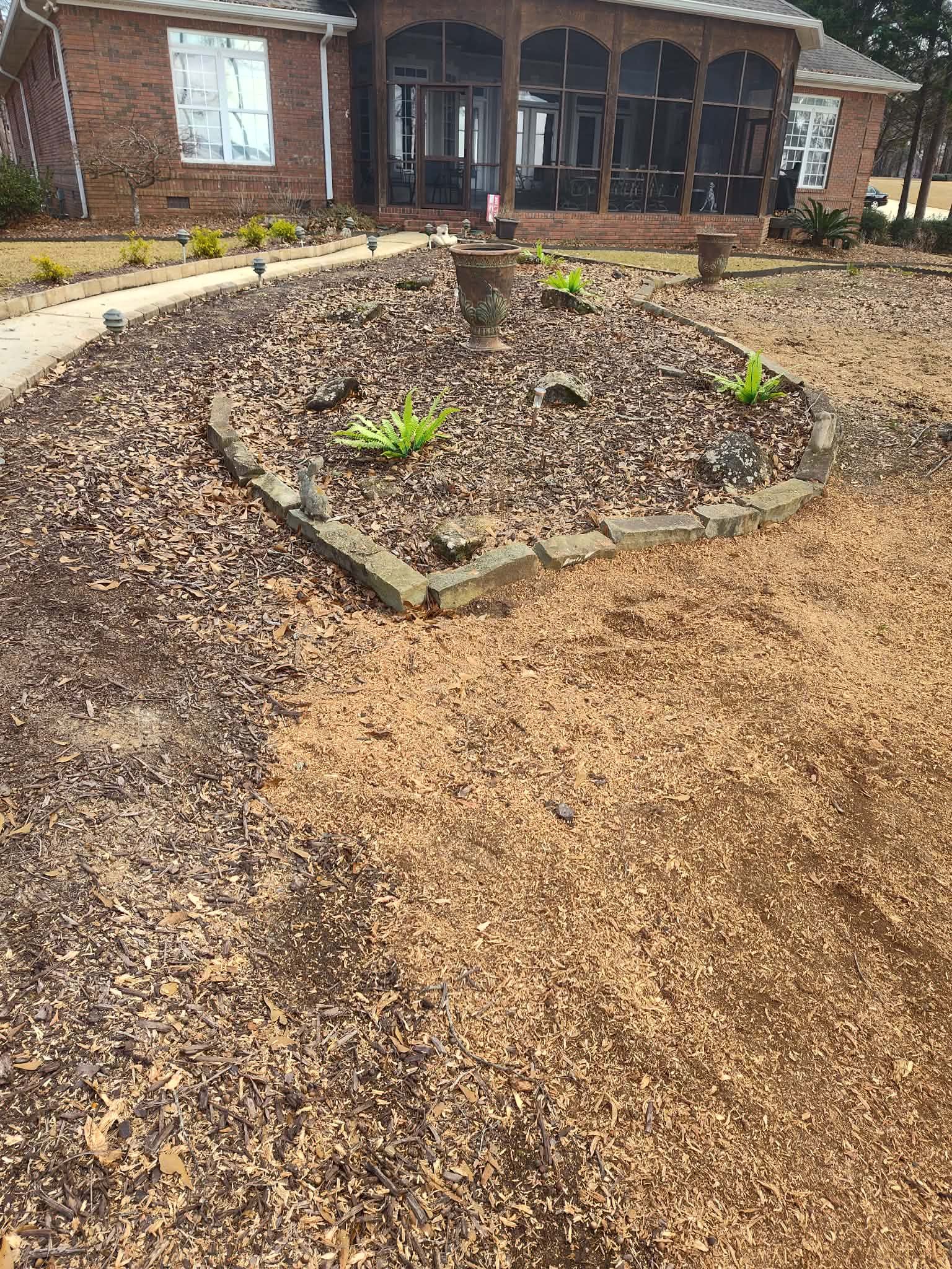 Flower bed in front yard with plants, outlined by stone, and surrounded by mulch and dry leaves.