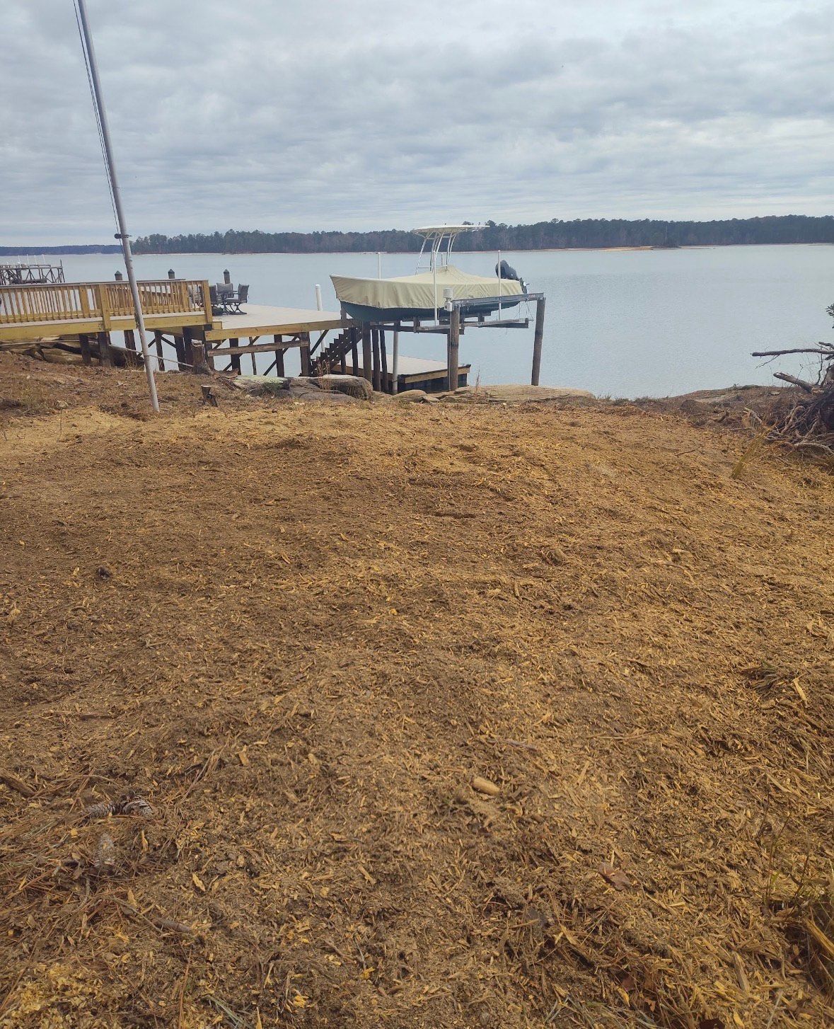 Pile of wood chips in front of a dock with a boat on a lift, overlooking a lake.