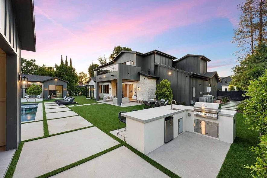 A modern backyard with a pool, a grassy lawn, stone patio, and an outdoor kitchen area with a grill at sunset.
