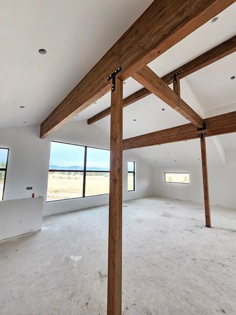 Interior of a room under construction with exposed wooden beams, white walls, and large windows.