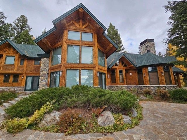 Log cabin house with stone accents, large windows, and green roof, surrounded by landscaping.