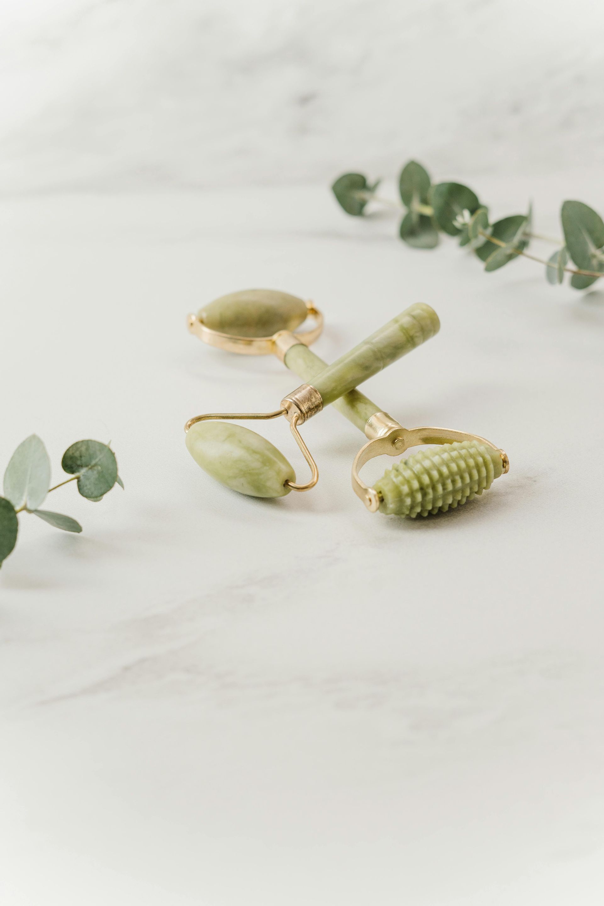 A green jade roller is sitting on a white table next to eucalyptus leaves.