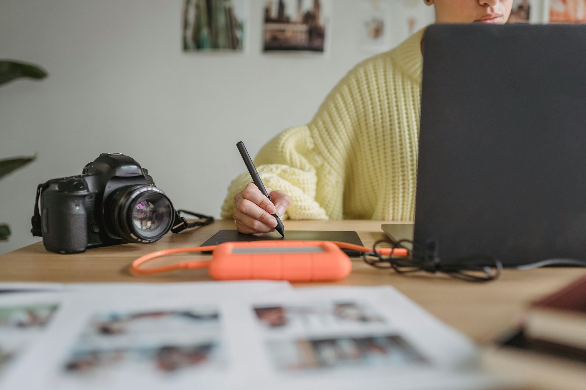Person writing beside a camera, orange external drive, and laptop on a desk