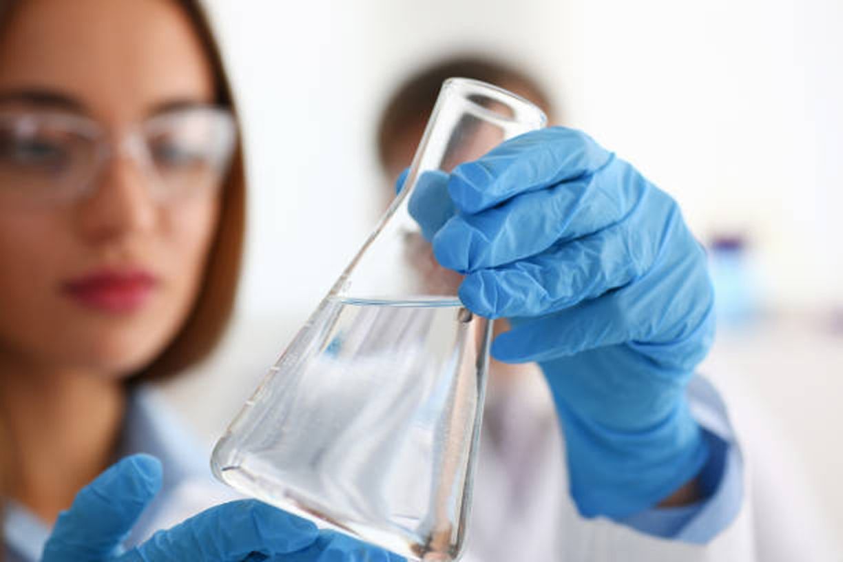 Scientist wearing blue gloves examines a transparent liquid in a conical flask, inside a lab.