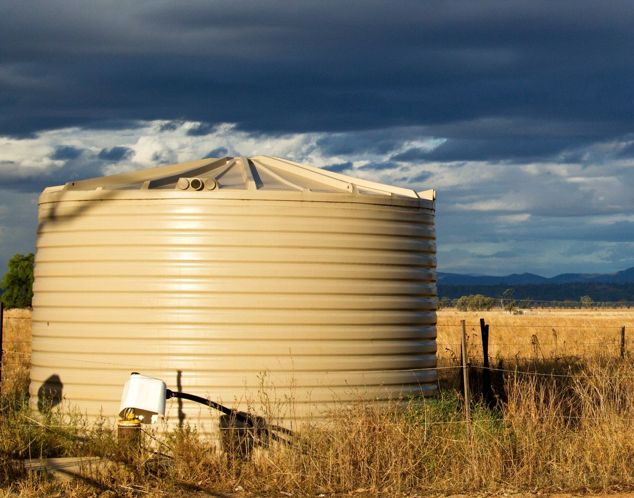 Large tan water tank in a field under a cloudy, dramatic sky.