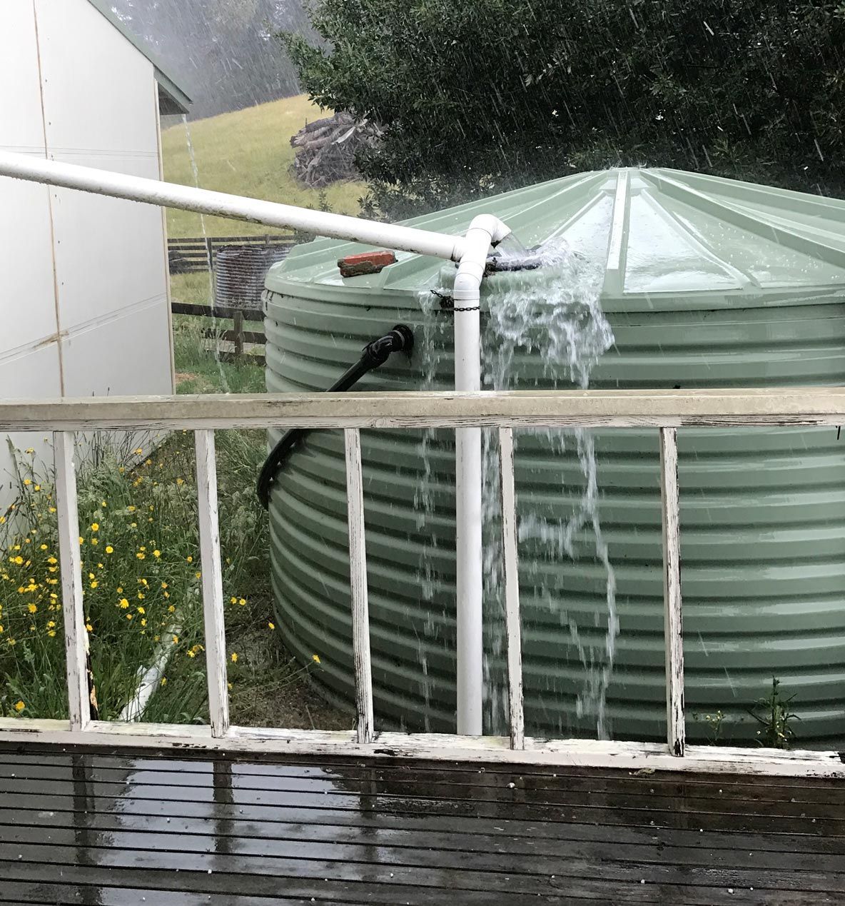Rainwater overflowing from a large green water tank with white piping, next to a porch.