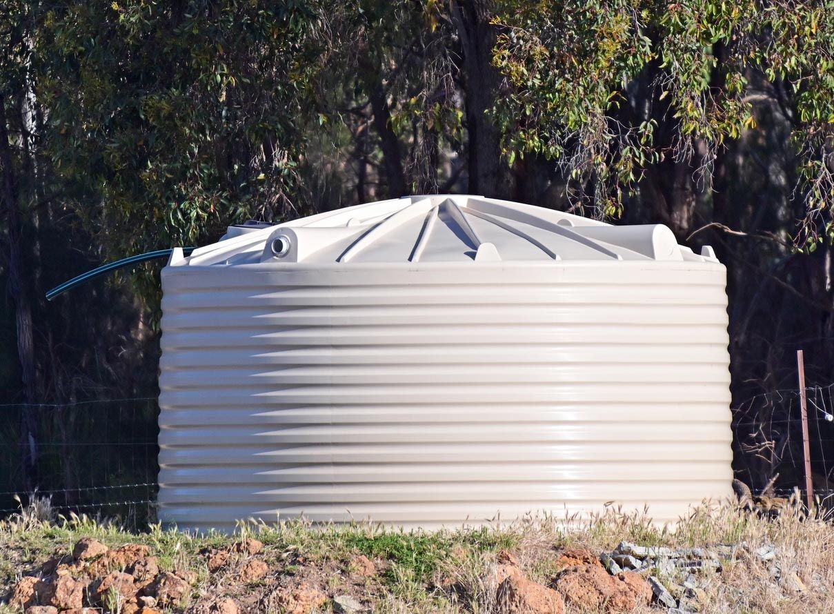 Tan corrugated water tank in a grassy area, trees in the background.