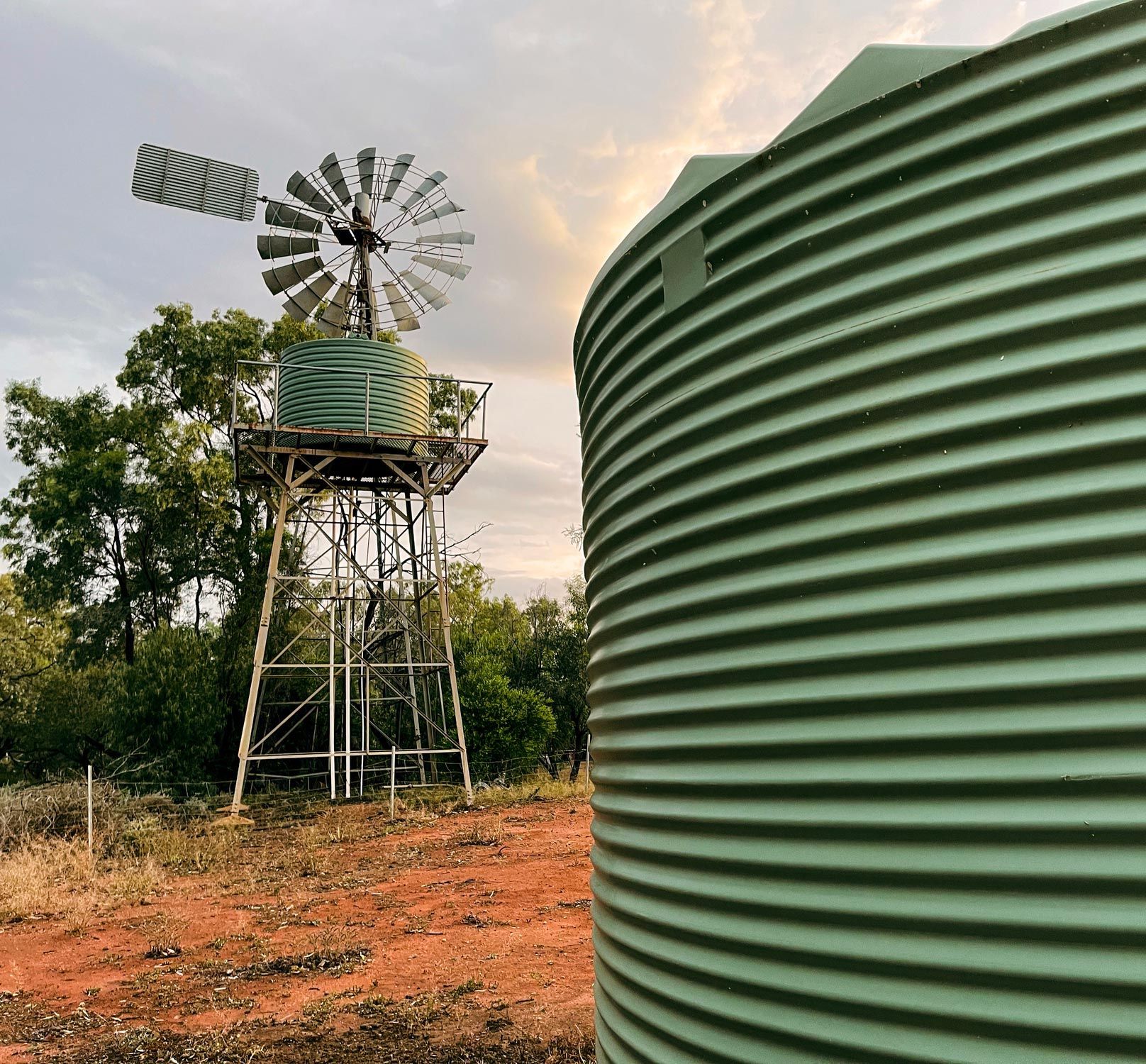 Windmill and water tank on reddish soil, with trees and a cloudy sky.