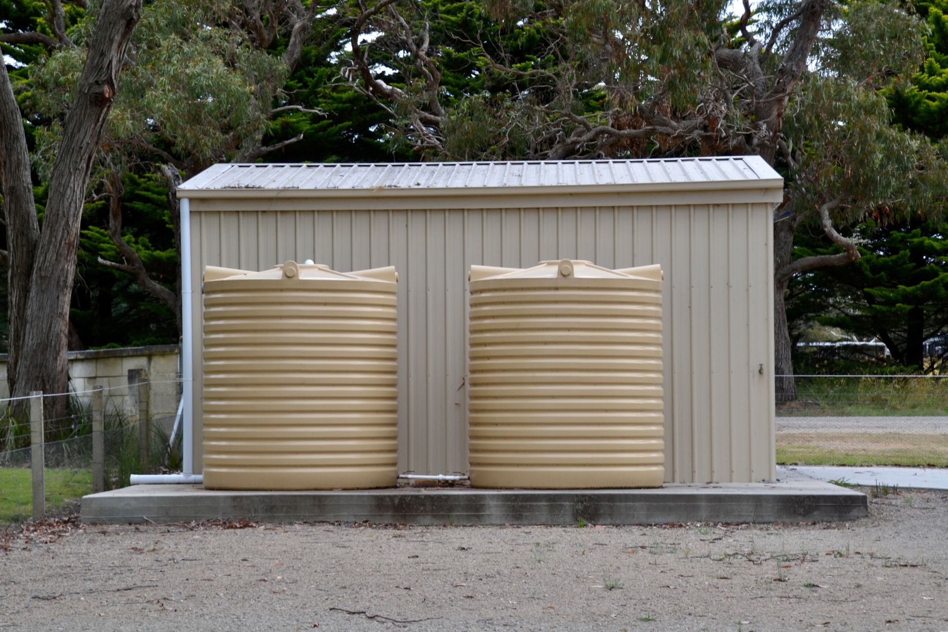 Two beige water tanks in front of a tan shed with a silver roof, set on a concrete pad.